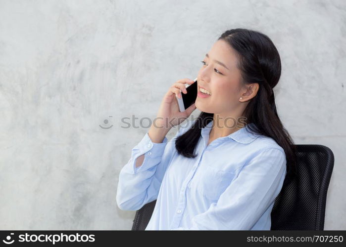 Beautiful young asian woman talking smart mobile phone and smile sitting on cement background, freelancer female calling telephone, communication concept.