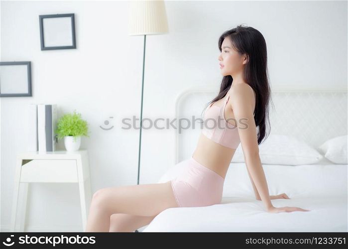 Beautiful young asian woman wake up and sitting and looking windows in the morning with cozy, sexy girl in underwear smiling relax and resting in the vacation with comfort, lifestyle concept.