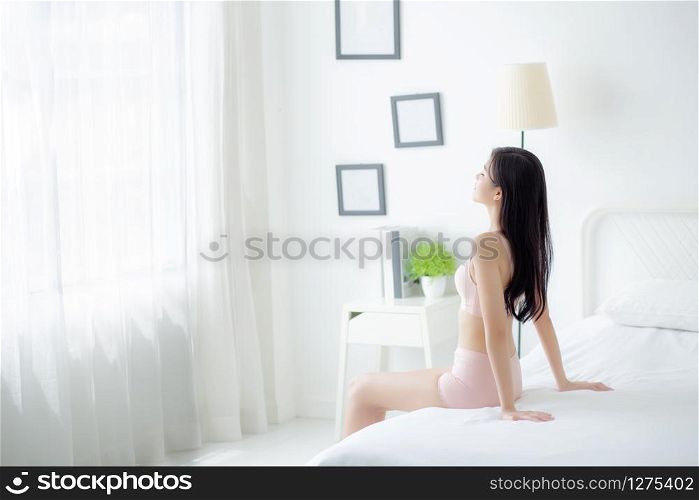 Beautiful young asian woman wake up and sitting and looking windows in the morning with cozy, sexy girl in underwear smiling relax and resting in the vacation with comfort, lifestyle concept.