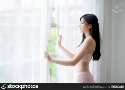 Beautiful young asian woman wake up and walking to window with open curtain in the morning with cozy, sexy girl in underwear smiling relax and resting in the vacation with happy, lifestyle concept.