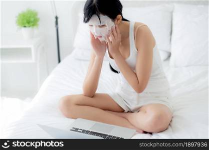 Beautiful young asian woman with sheet facial mask sitting working on laptop computer on bed at bedroom, beauty girl applying face mask for skincare of wrinkle at home, skin care, health and wellness.