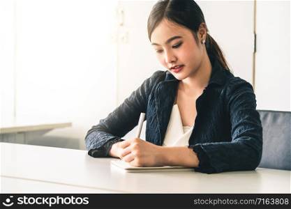 Beautiful young Asian woman writes letter on notebook while sitting at office desk. Content writer and secretary job concept.