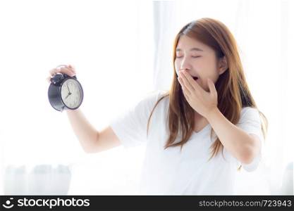 Beautiful young asian woman yawn wake up in morning annoyed alarm clock holding hand, standing hurry wake late with appointment with curtain background on bedroom, lifestyle concept.
