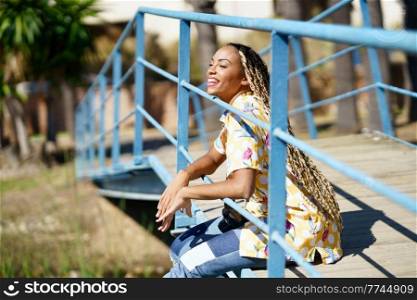 Beautiful young black woman combed with colorful braids sitting on an urban bridge with a camera on vacation.. Young black woman combed with braids sitting on an urban bridge with a camera on vacation
