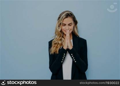 Beautiful young blond girl standing in praying pose folded palms together with her eyes closed, pleading and making wish come true, isolated over blue studio background with copy space. Beautiful young blond girl standing in praying pose folded palms together with her eyes closed