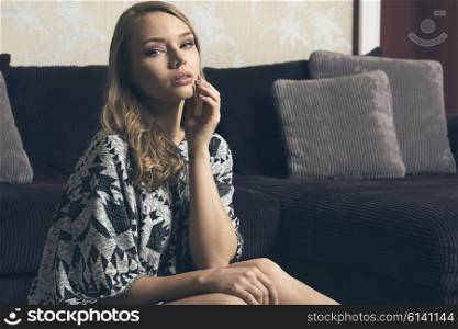 beautiful young blond lady , sitting on the floor near a big sofa , in her living room. she is looking in camera