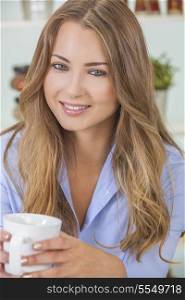 Beautiful young blond woman at home in her kitchen smiling and drinking a cup of tea or coffee