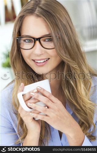 Beautiful young blond woman at home in her kitchen wearing geek glasses smiling and drinking a cup of tea or coffee