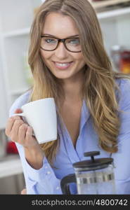 Beautiful young blond woman at home wearing glasses in her kitchen smiling and drinking a cup of tea or coffee