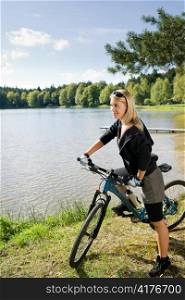 Beautiful young blond woman riding bicycle by lake summer day