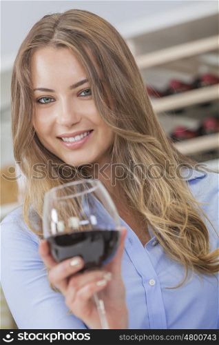 Beautiful young blond woman smiling and drinking a glass of red wine with bottles on a rack behind her