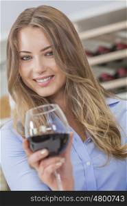 Beautiful young blond woman smiling and drinking a glass of red wine with bottles on a rack behind her