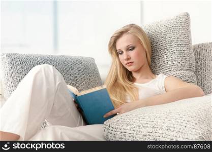 Beautiful Young Blonde Woman is Sitting on a Comfortable Sofa and is Reading a Book in a Blue Cover