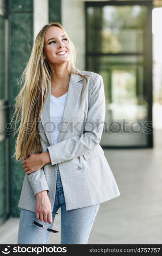 Beautiful young blonde woman smiling in urban background.. Beautiful young caucasian woman smiling in urban background. Blond girl wearing casual clothes in the street. Female with elegant jacket and blue jeans