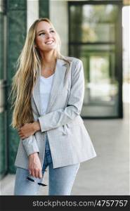 Beautiful young blonde woman smiling in urban background.. Beautiful young caucasian woman smiling in urban background. Blond girl wearing casual clothes in the street. Female with elegant jacket and blue jeans