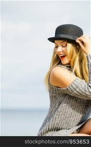 Beautiful young blonde woman wearing fedora hat, long warm sweater jumper posing next to sea beach.. Woman wearing fedora and jumper outdoor