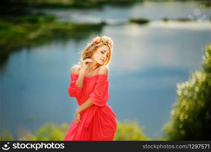 beautiful young blonde woman with a flower wreath on her head. Beauty girl with flowers hairstyle in a red long dress near the lake. Fashion photo, copy space.. beautiful young blonde woman with a flower wreath on her head. Beauty girl with flowers hairstyle in a red long dress near the lake. Fashion photo, copy space