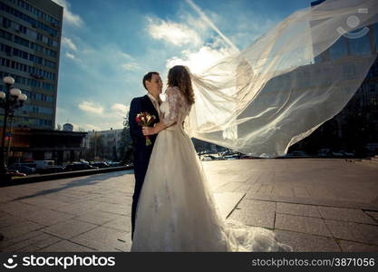 Beautiful young bride with long veil hugging with groom on street at windy day