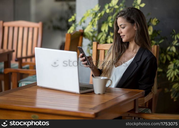 Beautiful young brunette in formal outfit sitting with coffee and laptop at table in hotel cafeteria using smartphone. Stylish businesswoman working in cafeteria