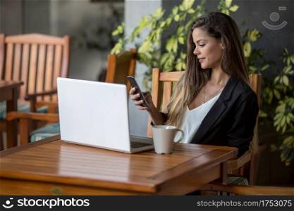 Beautiful young brunette in formal outfit sitting with coffee and laptop at table in hotel cafeteria using smartphone. Stylish businesswoman working in cafeteria