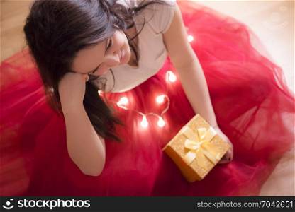 Beautiful young brunette woman, dressed in a red tutu, sitting on the floor, surrounded by Christmas lights and holding a gift box with a tied bow.