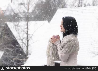 Beautiful young brunette woman in a cozy sweater and a shawl, holding a cup of hot drink, enjoying the falling snowflakes with her eyes closed.