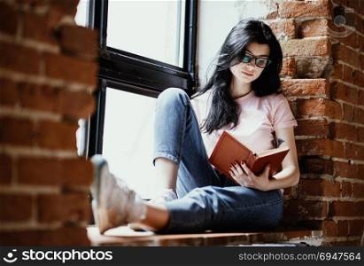 Beautiful young brunette woman reading book near window at home.. Beautiful young brunette woman reading book near window at home