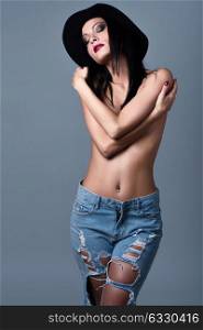 Beautiful young brunette woman wearing broken blue jeans and hat. Beautiful topless girl with make up closed eyes and crossed arms. Studio shot.