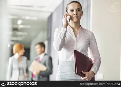 Beautiful young businesswoman on call with colleagues in background at office corridor