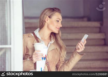 Beautiful young businesswoman texting and drinking coffee, working on the smartphone, modern computer technologies in business life