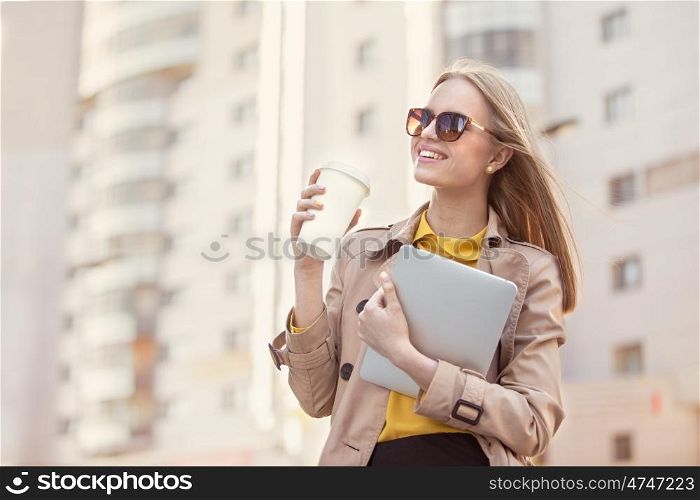 Beautiful young businesswoman with a disposable coffee cup, drinking coffee, and holding tablet in her hands against urban city background.