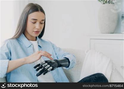 Beautiful young caucasian disabled girl turns on her bionic prosthetic arm. Woman with disability touches sensory robotic hand, artificial limb. People with disabilities, medical high tech. Copy space. Young disabled girl turns on her bionic prosthetic arm. People with disabilities, medical high tech