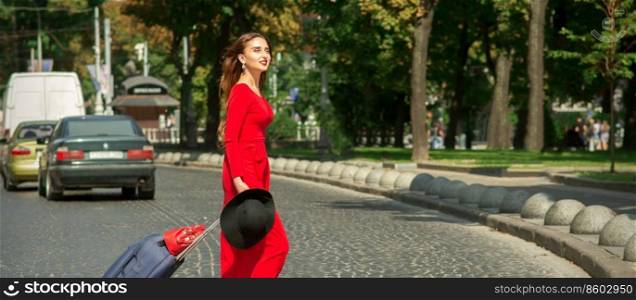 Beautiful young caucasian tourist woman with a suitcase in red long dress crosses the road through a crosswalk on the city street outdoors. Woman crosses road through crosswalk
