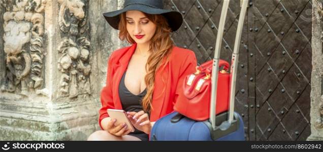 Beautiful young caucasian travel woman in a black hat with suitcase sitting and looking in a smartphone on the old city street. Woman looking in a smartphone