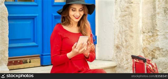 Beautiful young caucasian traveling woman in black hat looking on the smartphone smiling and sitting on stairs at the door outdoors. Woman looking on the smartphone