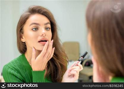 Beautiful young caucasian woman applies lipstick on her lips with her finger looking in mirror. Woman applies lipstick on her lips