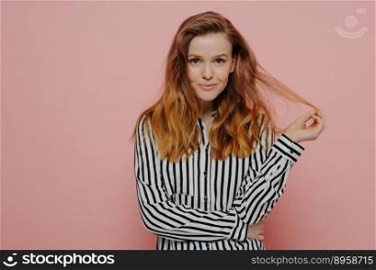 Beautiful young caucasian woman dressed in casual clothes playing with hair and looking at camera, cute female without makeup in shirt posing isolated on pink background. Studio shot. Young playful woman dressed in casual clothes playing with hair