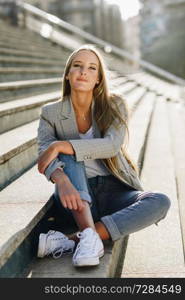 Beautiful young caucasian woman in urban background. Blond girl with elegant jacket and blue jeans sitting on stairs.