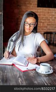 Beautiful young caucasian woman writes notes to notepad holding smartphone in her hand wearing glasses in cafe. Young woman writes notes to notepad