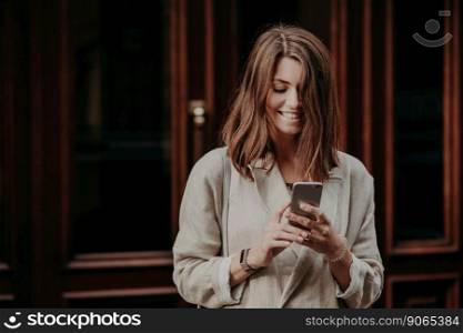 Beautiful young cheerful woman recieves notification message, gets advertising offer, has trendy haircut, dressed in fashionable clothes, stands outdoor, walks to work, reads latest news in internet
