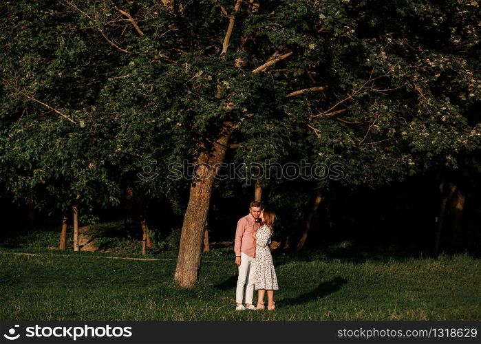 beautiful young couple in love hugging and smiling in the summer park. Stylish man in pink shirt and woman in dress are having fun together. love story. copy space.. beautiful young couple in love hugging and smiling in the summer park. Stylish man in pink shirt and woman in dress are having fun together. love story. copy space