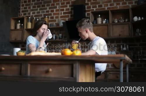 Beautiful young couple in love spending nice morning together in domestic kitchen. Handsome hipster drinking orange juice from mason jar and chatting with his cute girlfriend. Loving couple having breakfast in the morning.