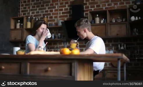 Beautiful young couple in love spending nice morning together in domestic kitchen. Handsome hipster drinking orange juice from mason jar and chatting with his cute girlfriend. Loving couple having breakfast in the morning.