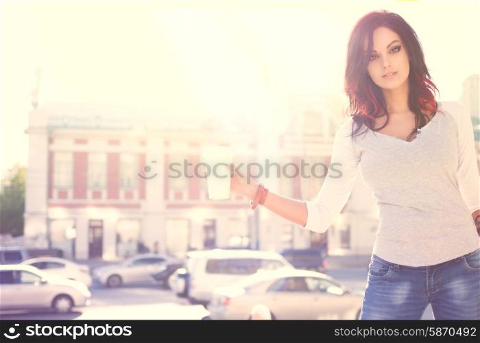 Beautiful young fashionable woman standing against urban city background holding a disposable takeaway cup with hot coffee.