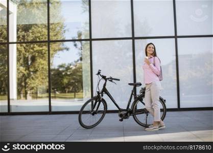 Beautiful young female cyclist drinks coffee from a cup by the electric bicycle in the urban environment
