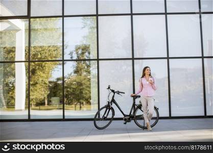 Beautiful young female cyclist drinks coffee from a cup by the electric bicycle in the urban environment