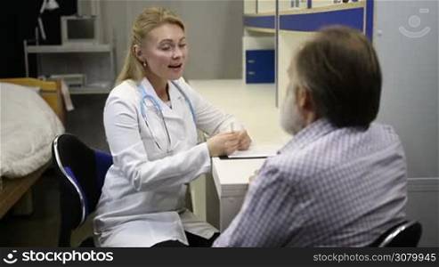 Beautiful young female doctor in lab coat asking senior patient for symptoms of illness during medical exam at clinic. Old male patient explaining his symptoms to attentive female physician at medical consultation