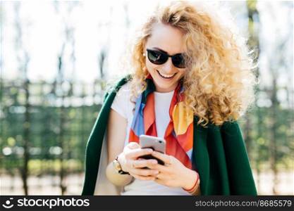 Beautiful young female in fashionable round shades texting messages to her friends via social networks while standing outside in green park enjoying good sunny weather. People, communication, lifestyle