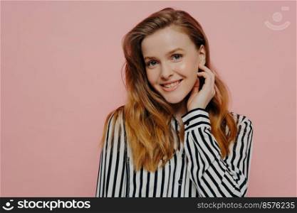 Beautiful young female in stripy black and white blouse with wavy ginger medium length hair touching face while posing over light pink studio background. Positive young woman smiling at camera posing against pink wall