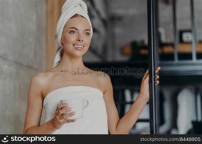 Beautiful young female looks thoughtfully into distance, drinks coffee or tea, poses with bare shoulders, wrapped in bath towel after showering, smiles pleasantly, has white teeth. Skin care concept